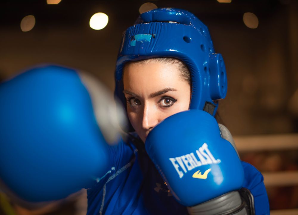 A female boxer wearing a blue headguard and gloves looks straight ahead as she throws a punch